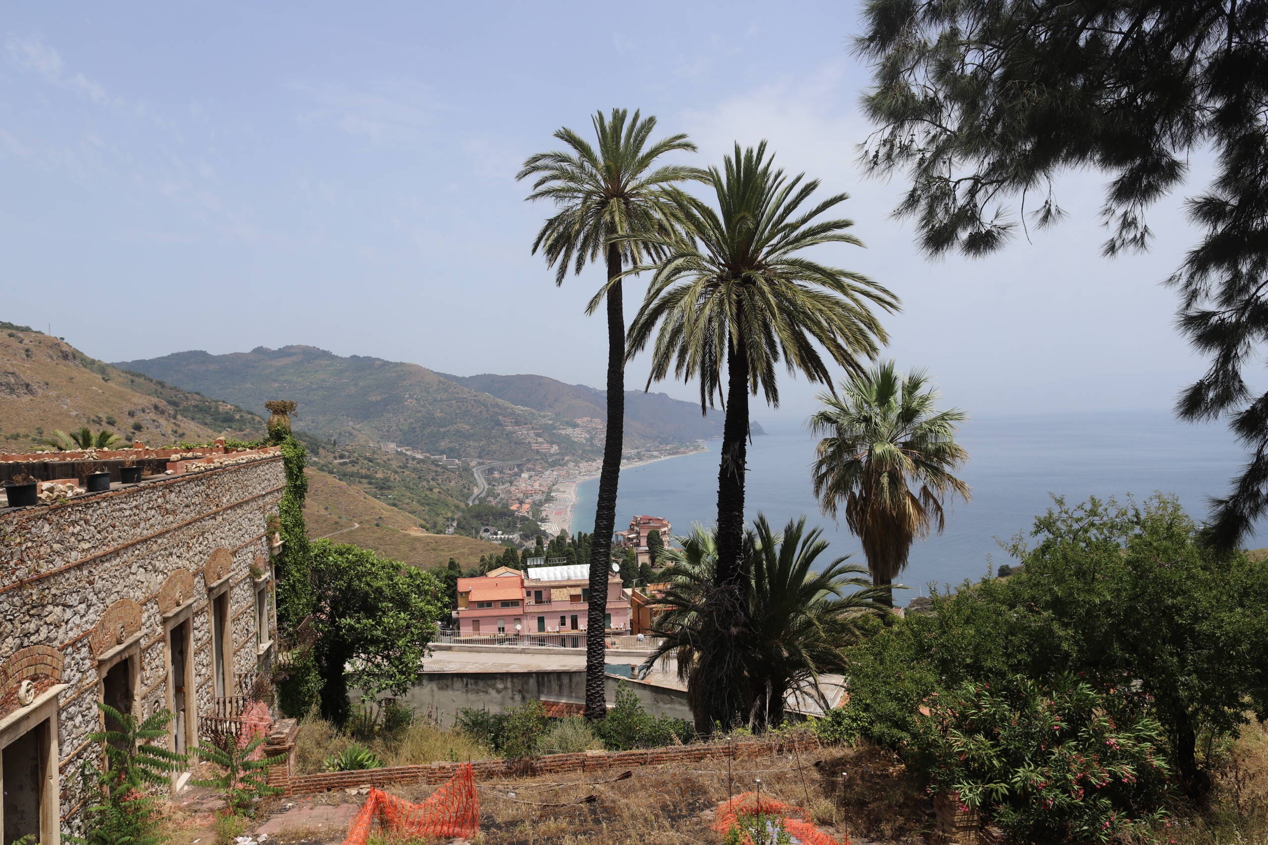 View from the upper hillside of Taormina, with date palms, a ruined stone building, and the Ionian coast curving toward Giardini Naxos in the distance