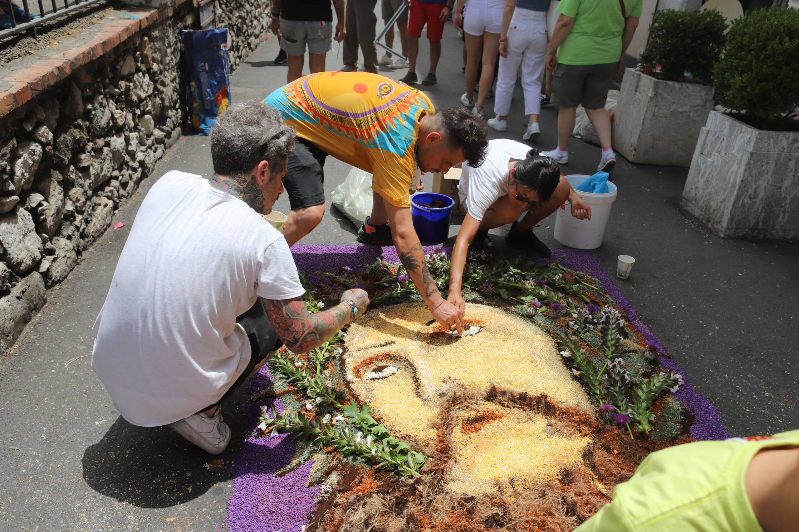 Three men kneeling on the street in Taormina, building an infiorata — a large figurative face rendered in colored sand, seeds, flowers, and herbs on the pavement