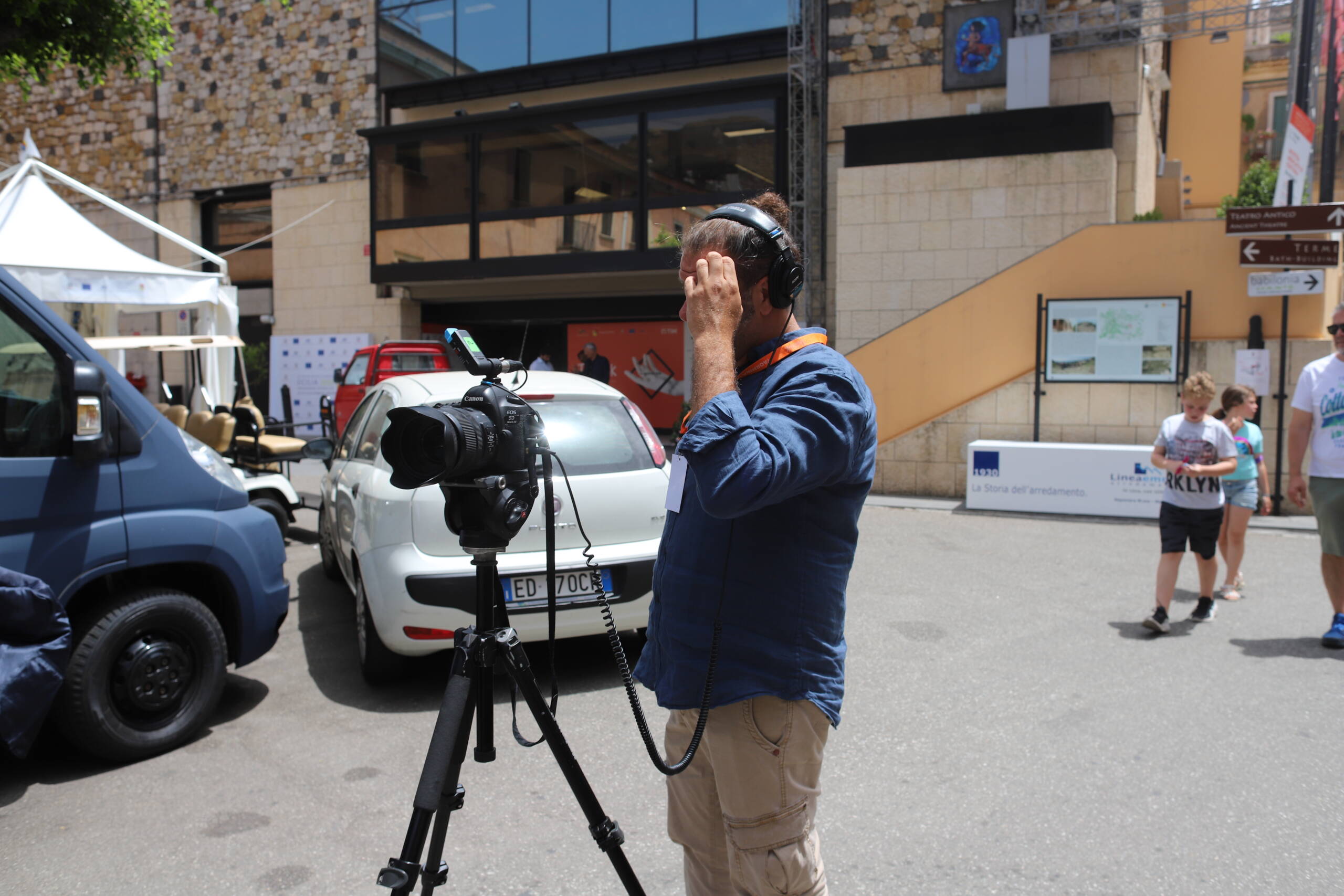 A cameraman with a Canon EOS on a tripod and headphones, shading his eyes in the midday glare outside a building near the Teatro Antico sign in Taormina