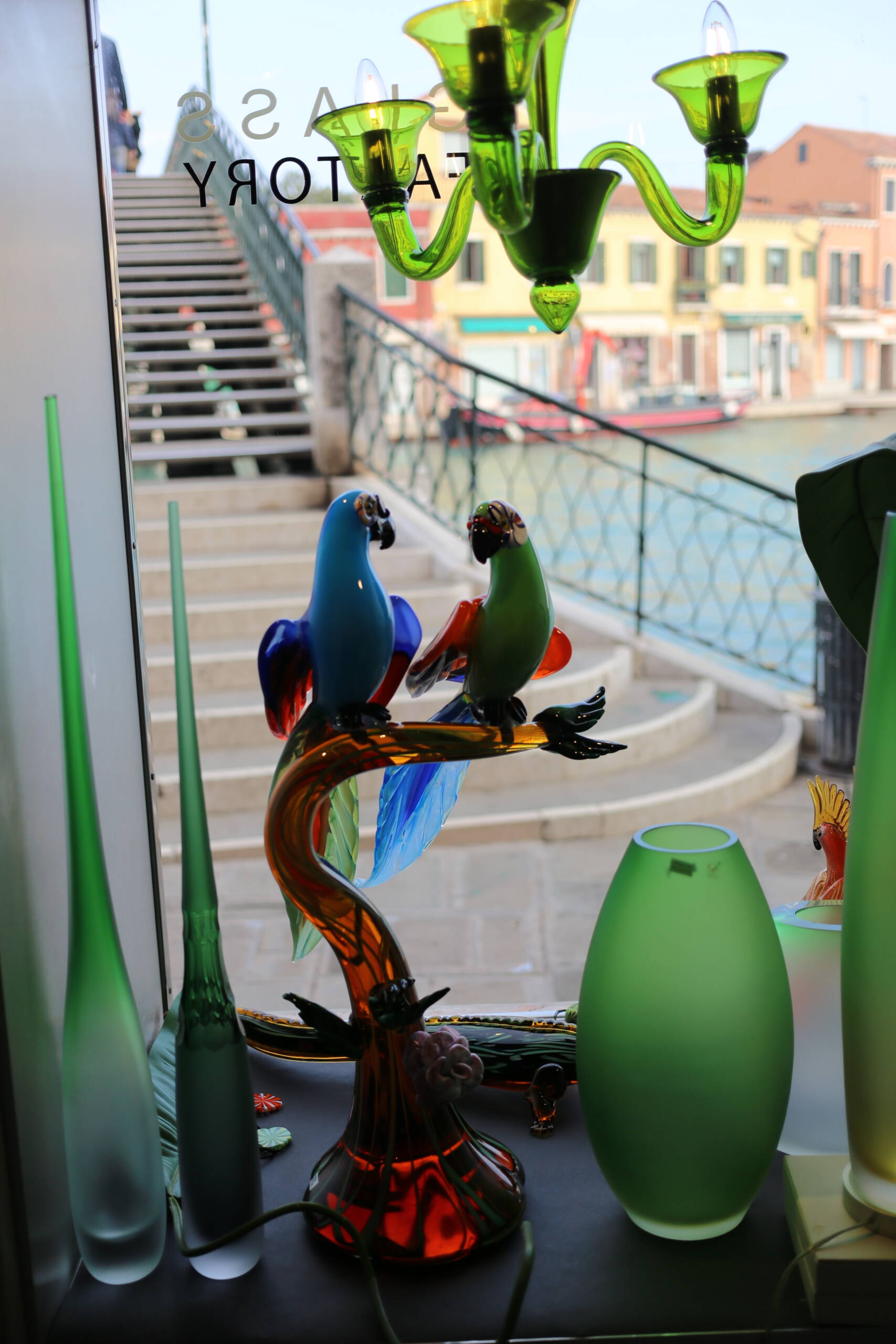 Blown-glass parrots and green chandelier in a Murano glass shop window with canal bridge visible behind