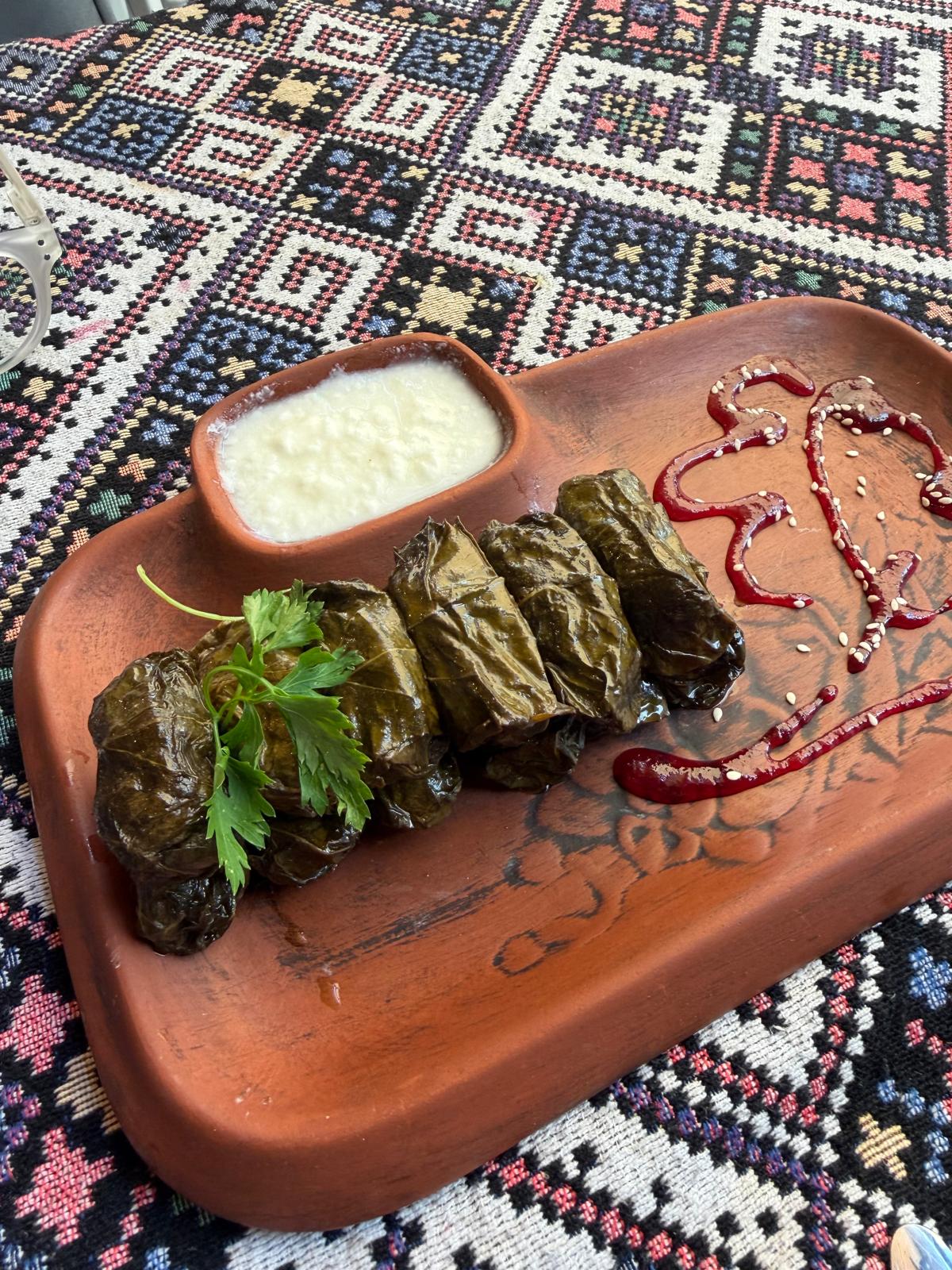 Yarpaq dolması served on a clay board with garlic yogurt and cornelian cherry sauce at Old Garden, Baku
Five vine-leaf rolls, garlic yogurt in the well of the board, a cornelian cherry glaze drawn across the wood.