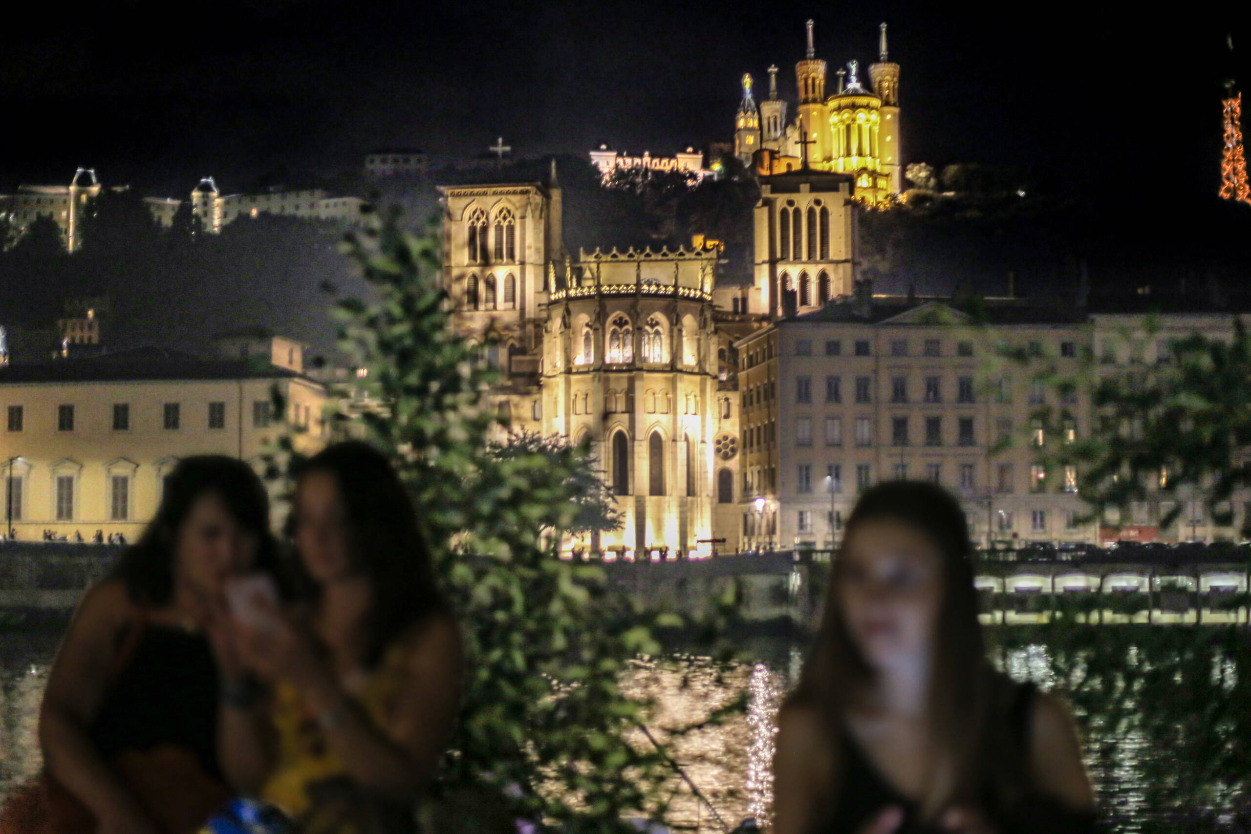 Lyon at night seen across the Saône, the Basilica of Notre-Dame de Fourvière lit gold on the hill and the Cathédrale Saint-Jean below, with three young women out of focus in the foreground looking at their phones.
