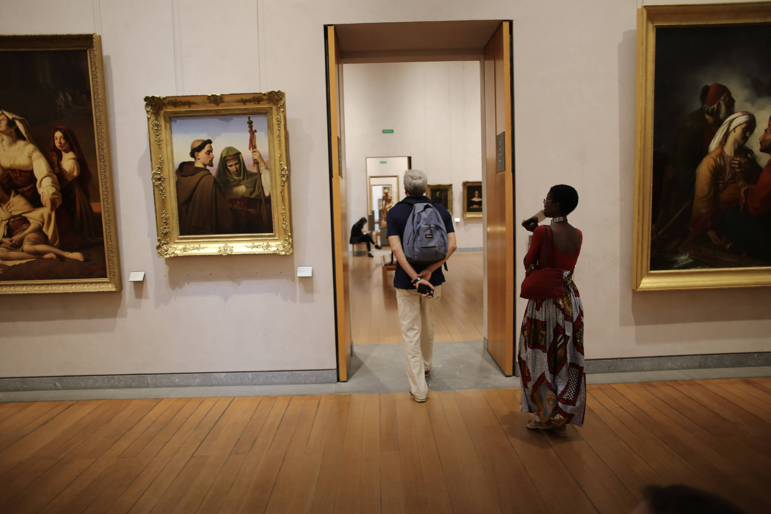 A nineteenth-century painting gallery with two visitors, one walking through an open doorway into a further room and one standing to the right looking at a canvas
