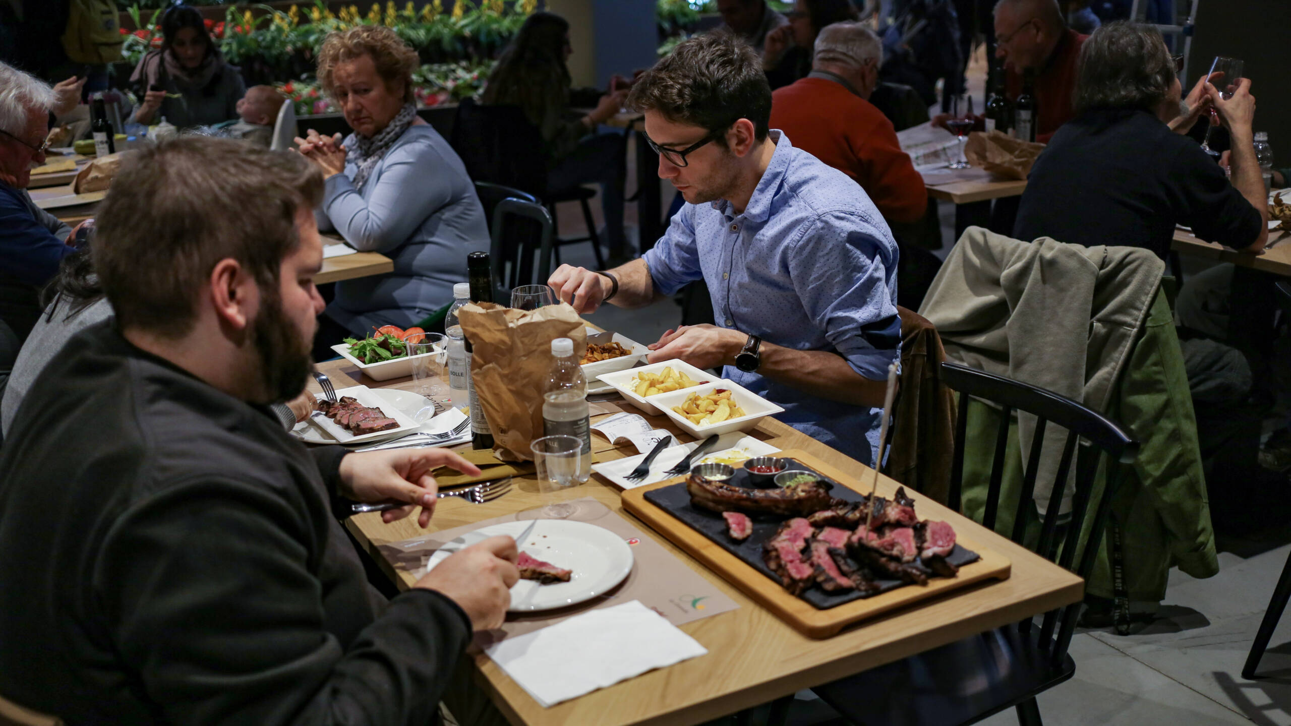 A crowded market-hall restaurant in Italy, sliced rare bistecca on a slate board with fries and a bone, diners at long wooden communal tables