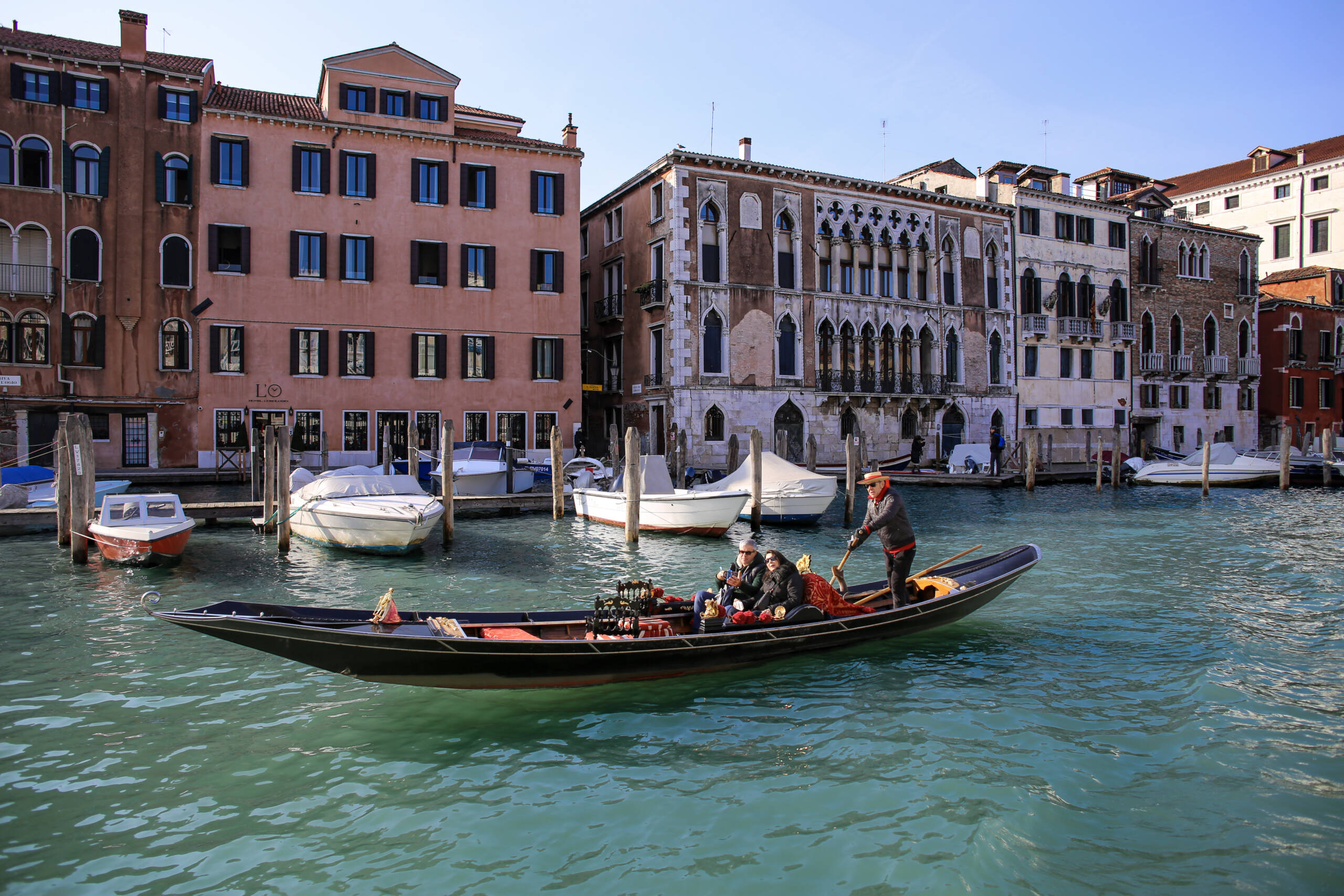 A gondola with two passengers on the Grand Canal in Venice on a clear winter day, moored motorboats along the quay and Gothic palazzo façades behind