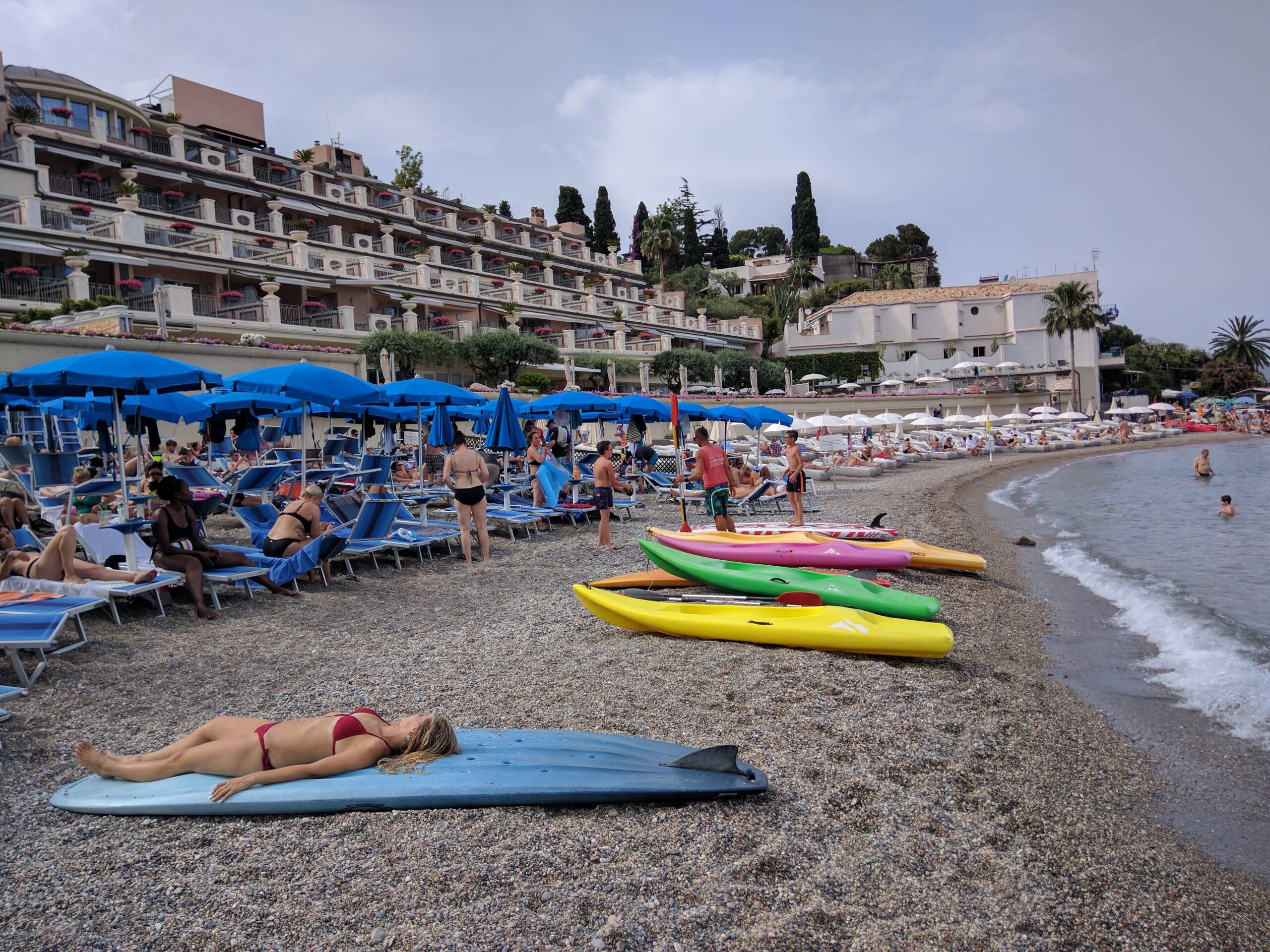 Crowded pebble beach at Mazzarò below Taormina, Sicily, with blue lido umbrellas and a terraced hotel built into the hillside