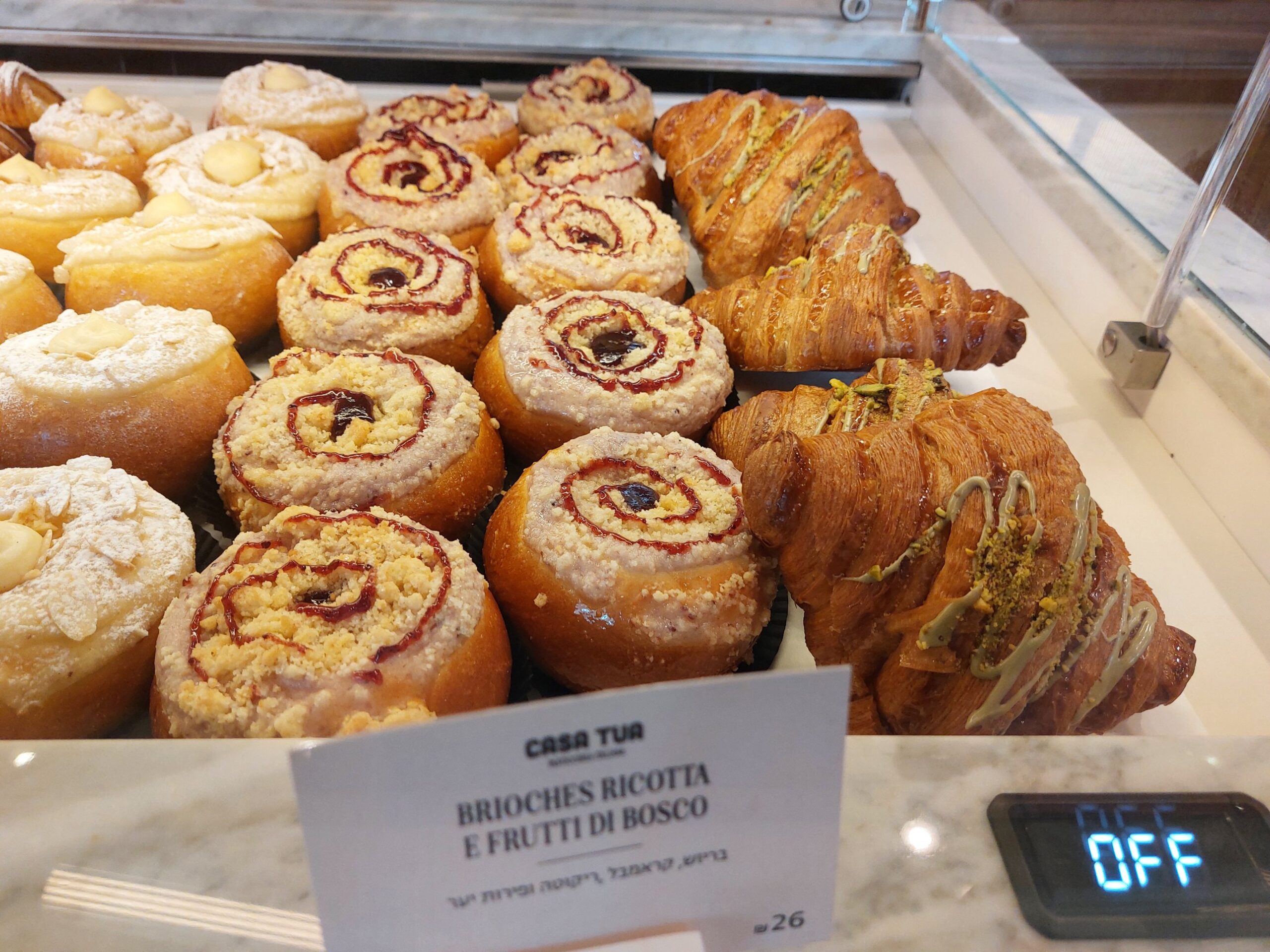 Ricotta and berry brioche with crumble and a red swirl, labelled Brioches Ricotta e Frutti di Bosco at 26 shekels, beside pistachio croissants