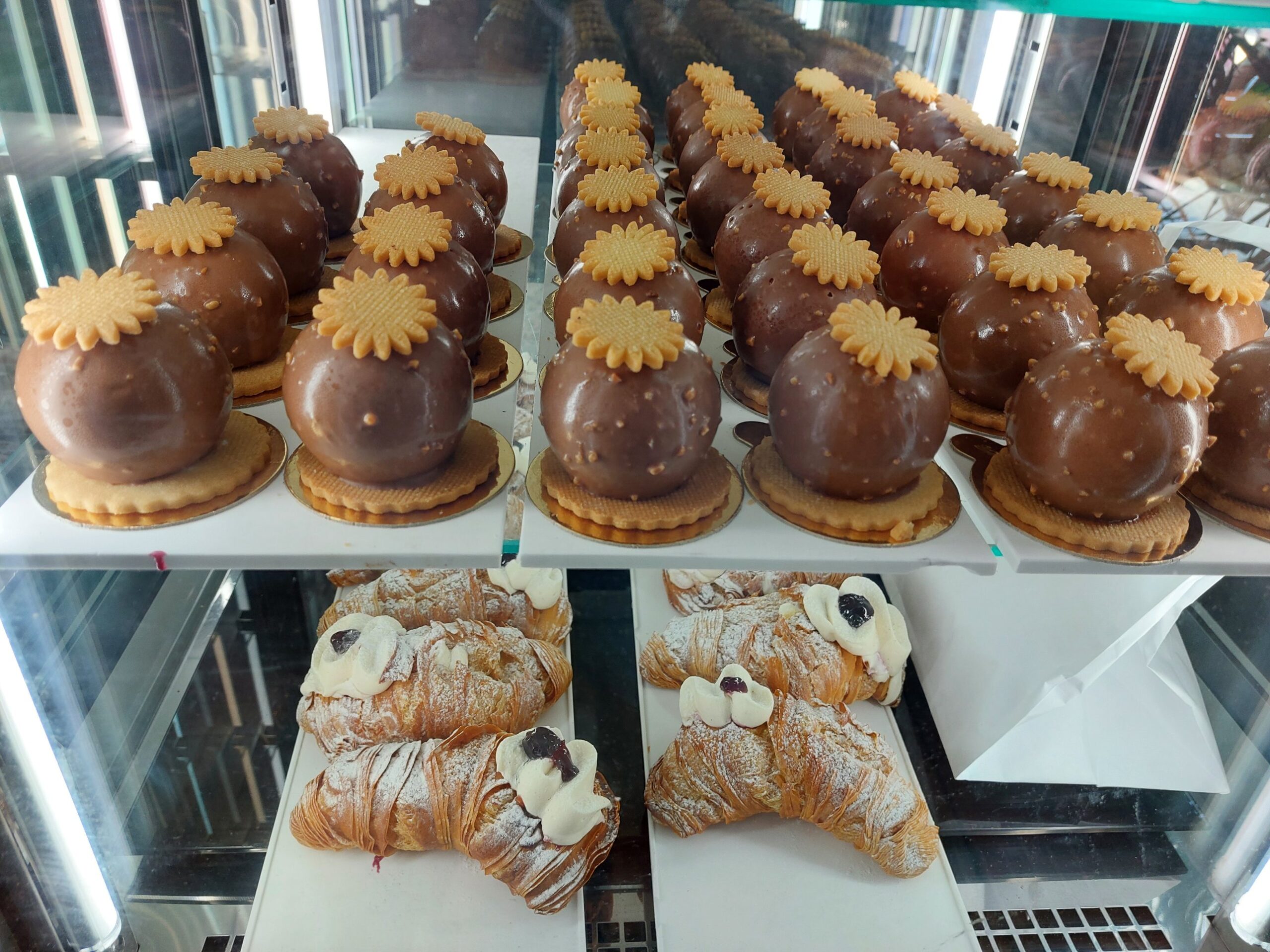Rows of glazed chocolate-hazelnut domes topped with biscuit flowers, above flaky cannoli filled with cream and cherry
