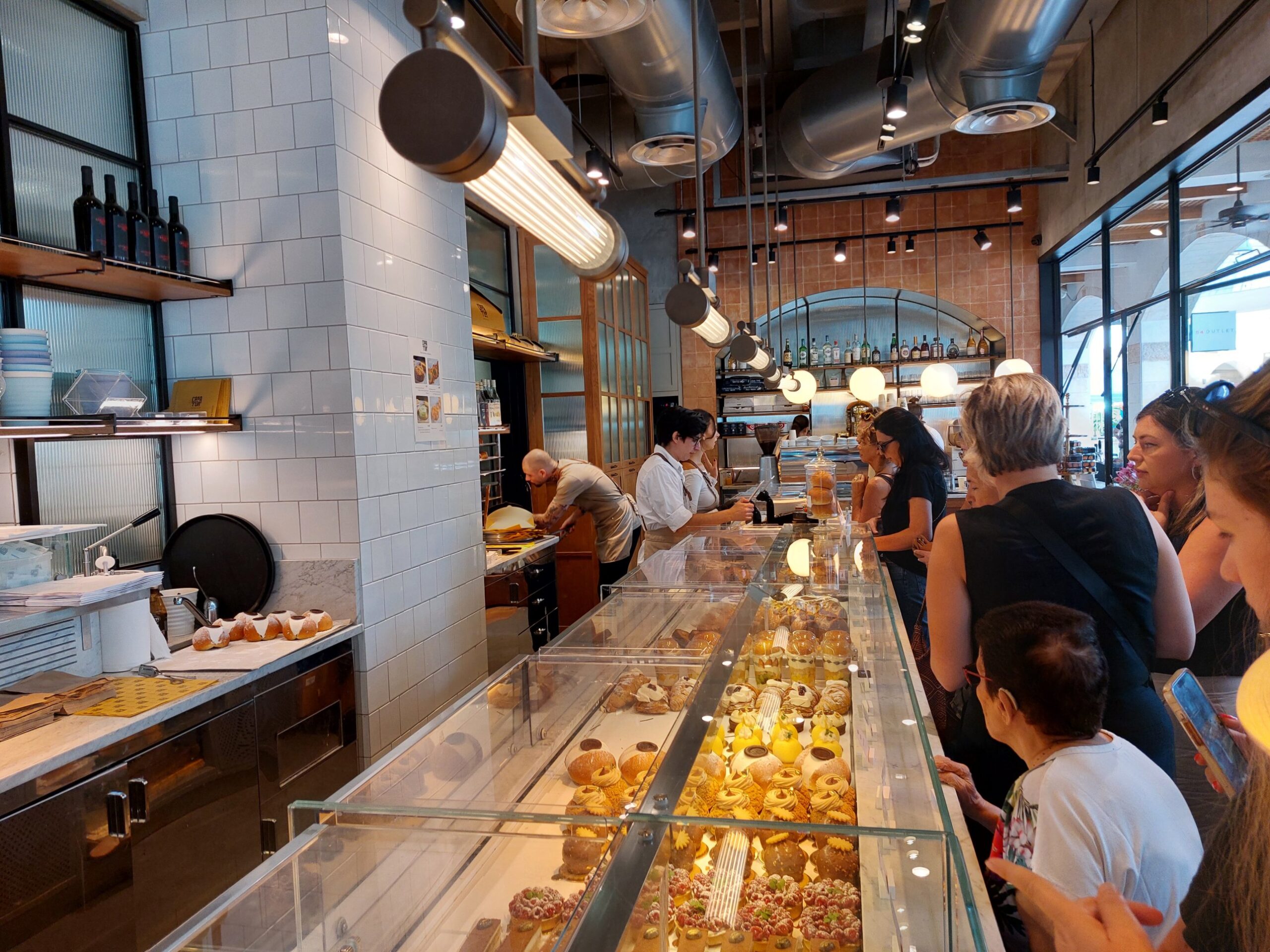 Inside Casa Tua in Herzliya: a long glass pastry counter, white tiled walls, staff at work and customers choosing pastries