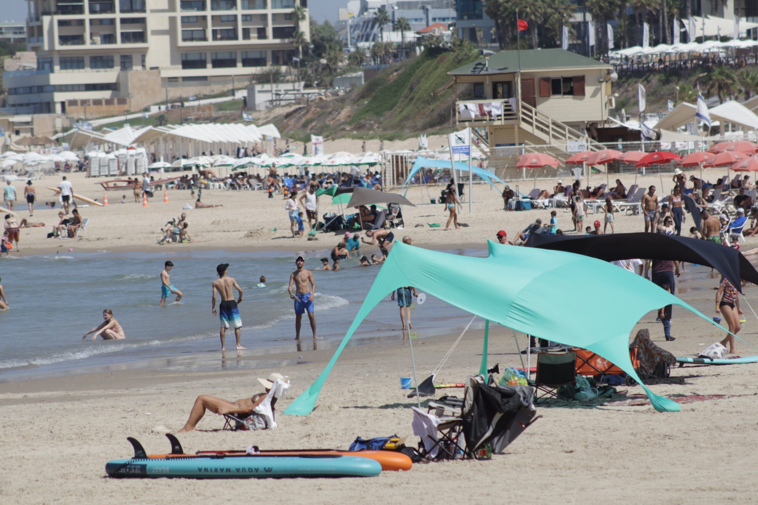 Herzliya beach with paddleboards on the sand, shade tents, red umbrellas and a wooden lifeguard tower