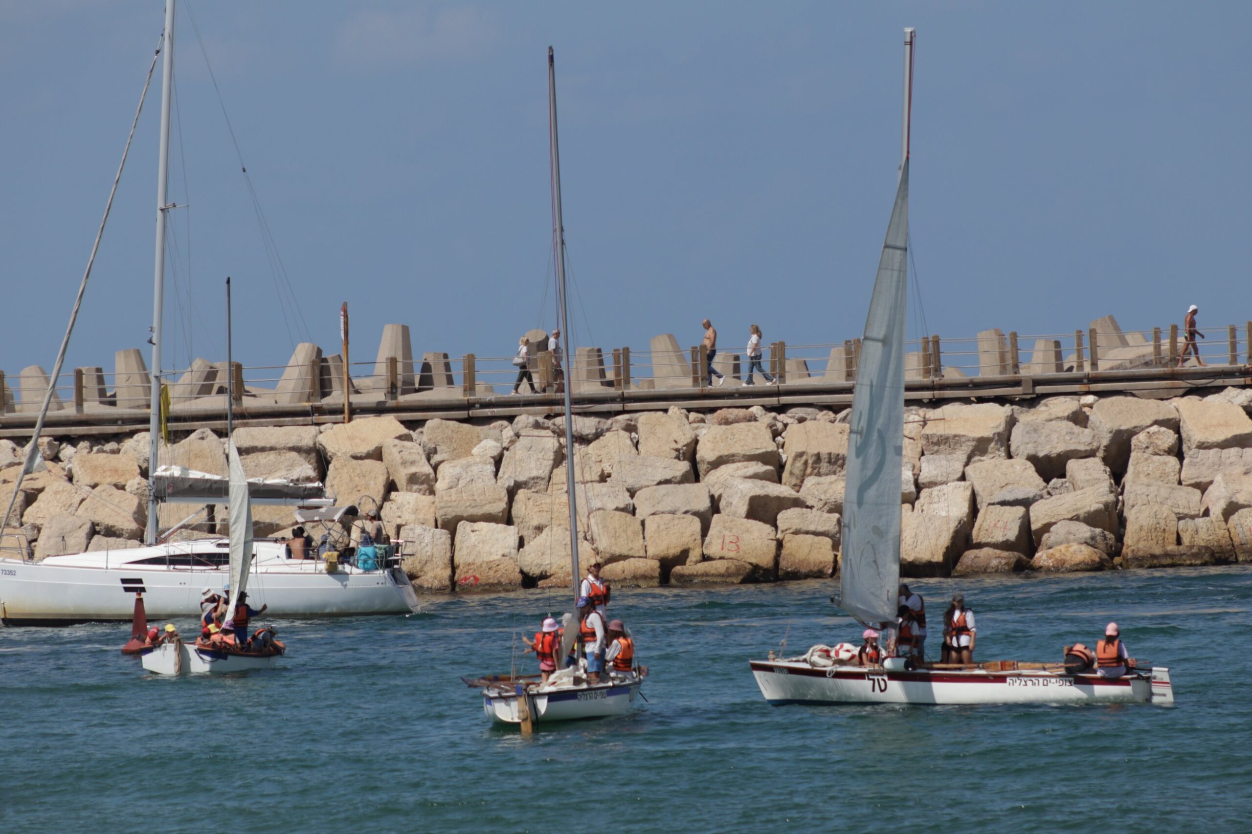 Sea scouts in life jackets sailing small boats past the stone breakwater of Herzliya Marina, with walkers on the promenade above